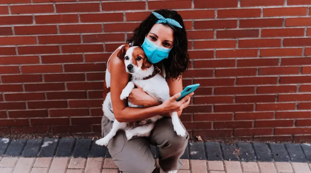 Women wearing a face mask holding her dog while using her phone