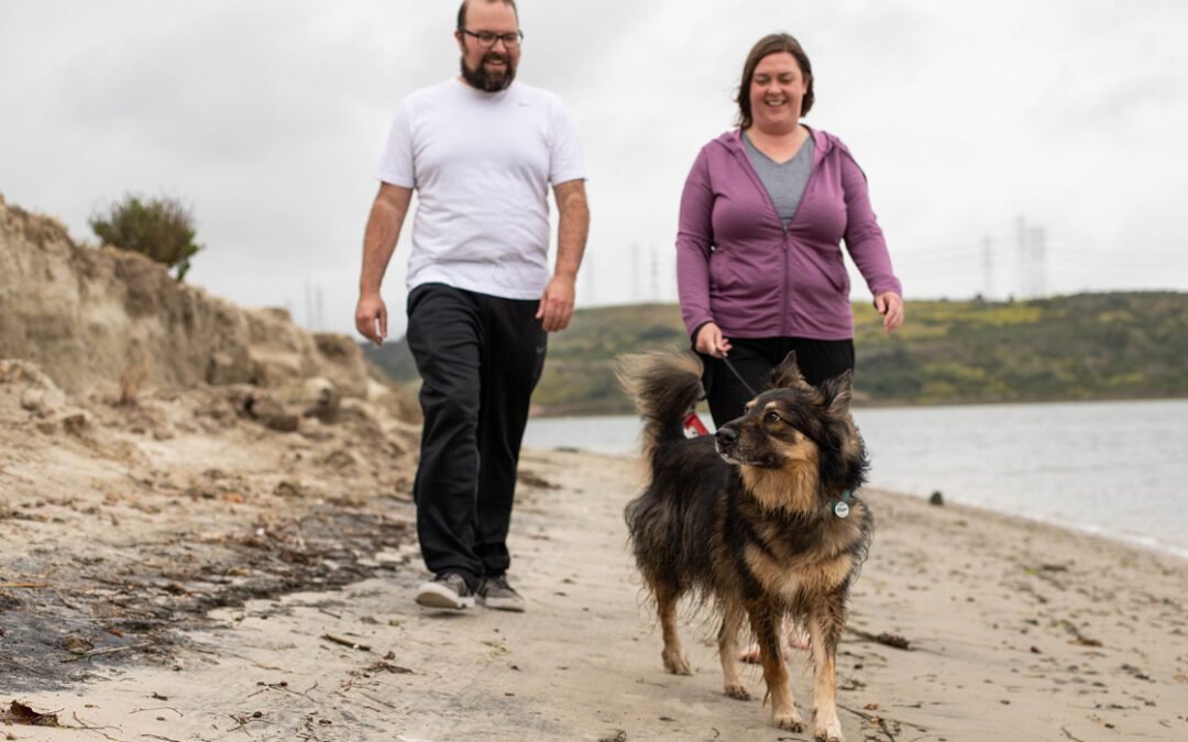 Male and female pet parent happily walking their dog in the sand