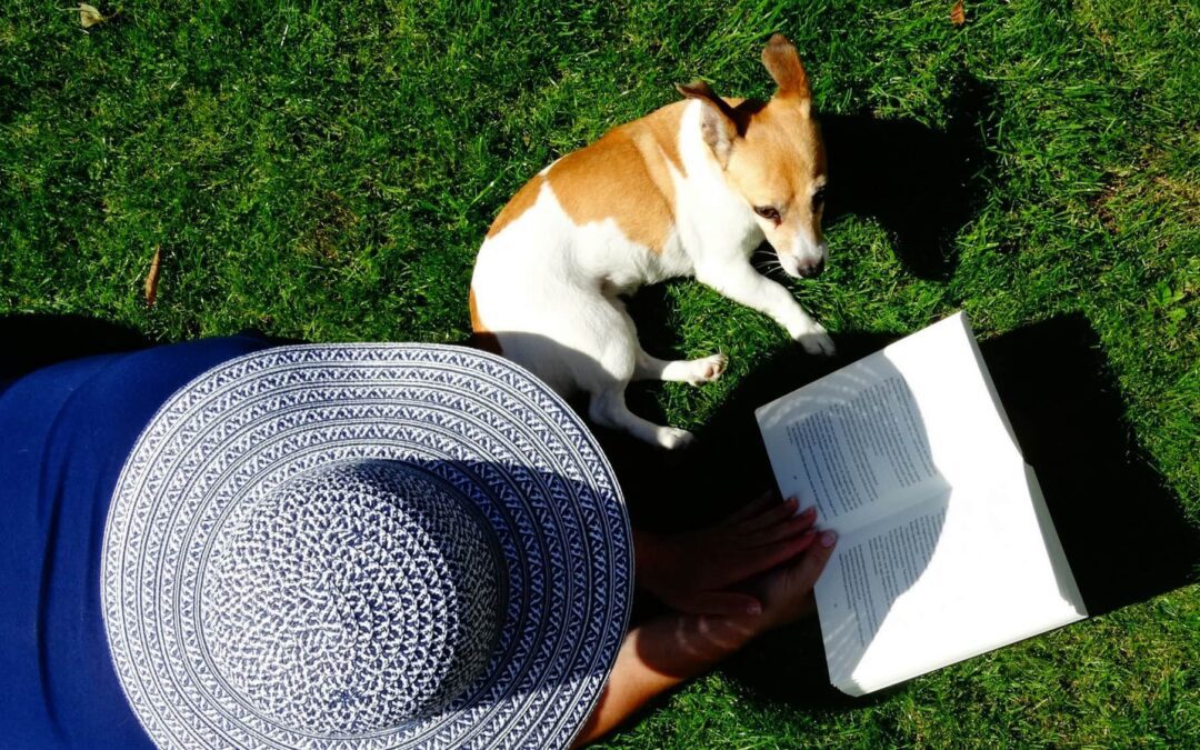 A woman laying in the grass wearing a hat and reading a book with their dog laying next to them